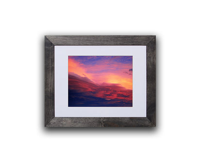 Nature photography print of a luminous blue, pink, yellow and red sunset shown with a black frame on a white wall called Purple by Loud Hue